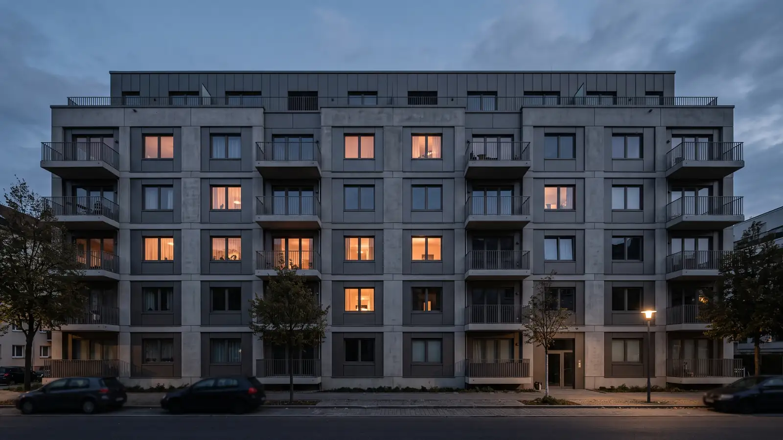 Fassade eines Mehrfamilienhauses in der Abenddämmerung, einzelne Fenster mit warmem Licht erleuchtet